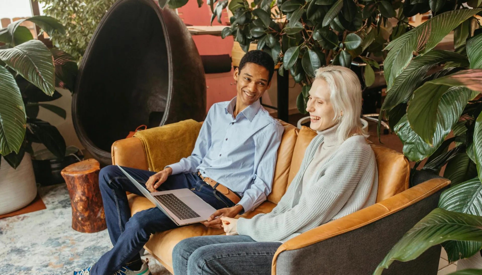 Two colleagues smiling and collaborating on a laptop in a cozy, plant-filled office setting.
