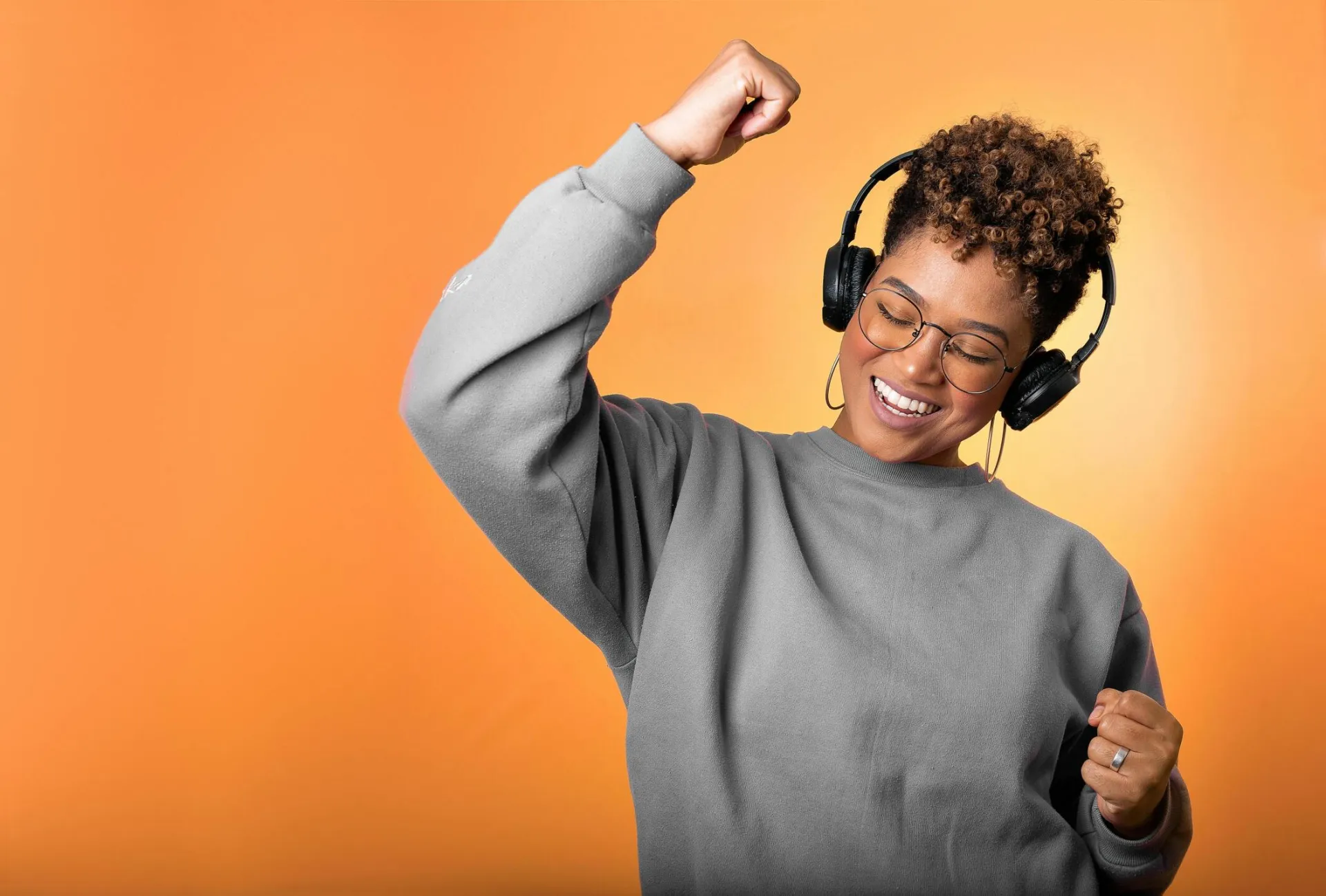 Smiling woman wearing headphones and eyeglasses, enjoying music on an orange background.