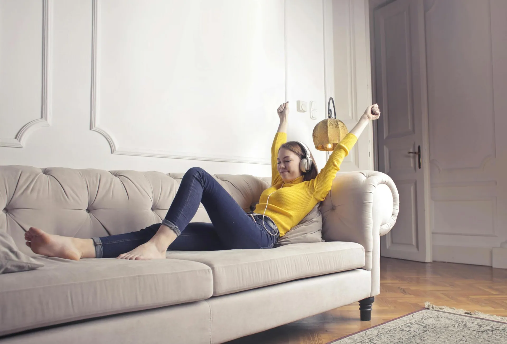 A woman in a yellow top relaxes on a sofa, enjoying music through headphones.