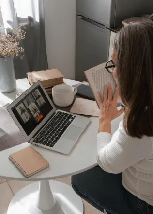 A woman participates in an online book club meeting using her laptop while holding a book at a home table.