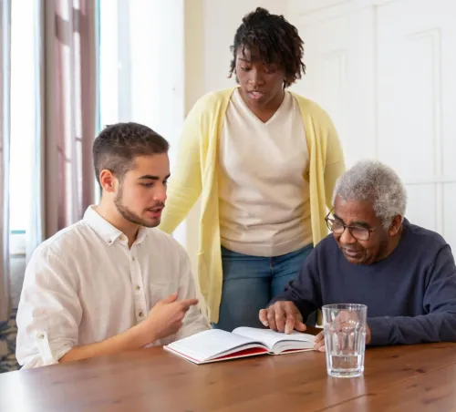 Young adults assisting an elderly man with reading in a nursing home.