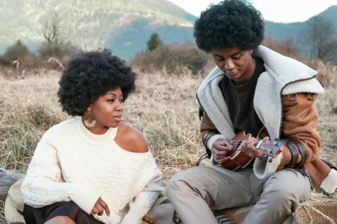 A stylish couple sits outdoors with a ukulele in a scenic grassy field.