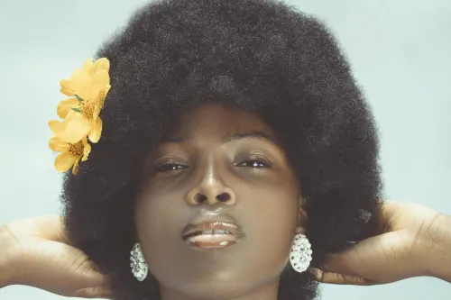 Close-up portrait of a woman with an afro, adorned with jewelry and a flower, posing confidently indoors.