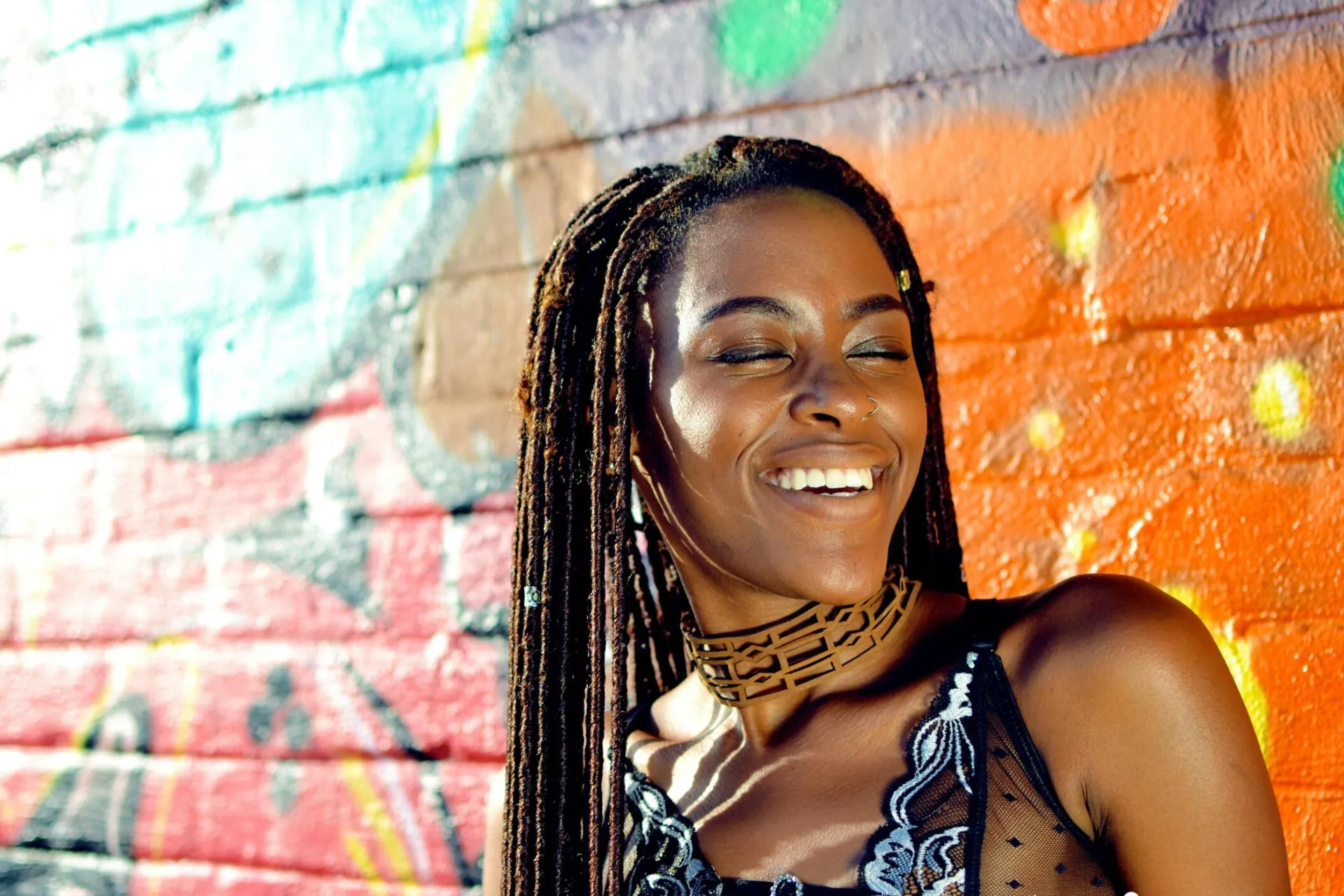 A smiling woman with locs in front of a colorful graffiti wall, exuding happiness.