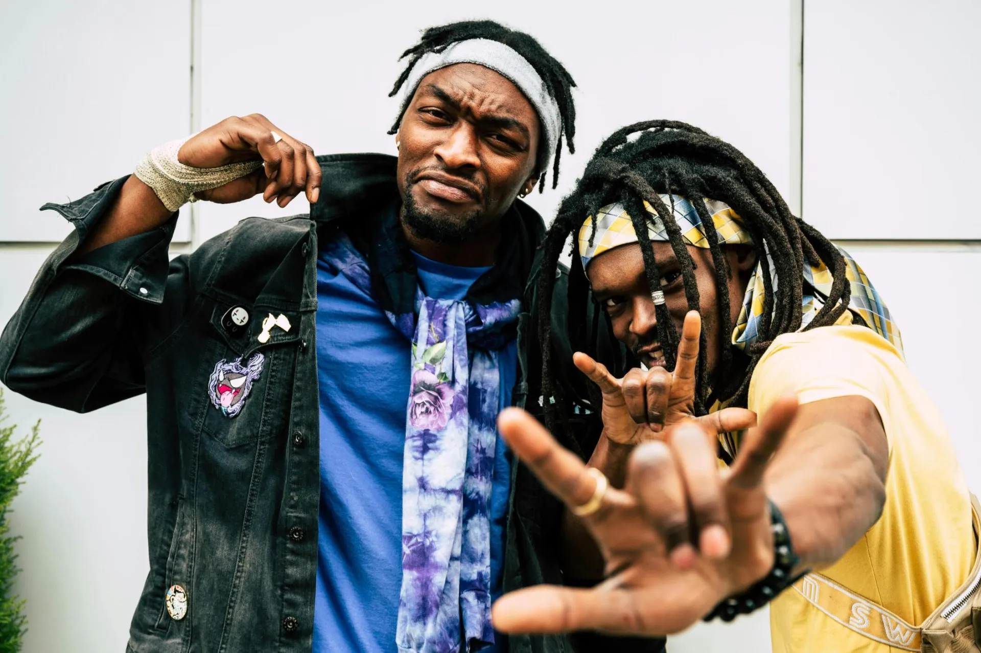 Smiling African American man with dreadlocks and headband showing sign of horns while frowning male friend pulling collar of coat and looking at camera