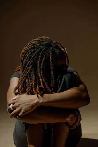 A woman with dreadlocks embraces herself in a thoughtful pose. Captured with warm tones, this portrait conveys introspection.