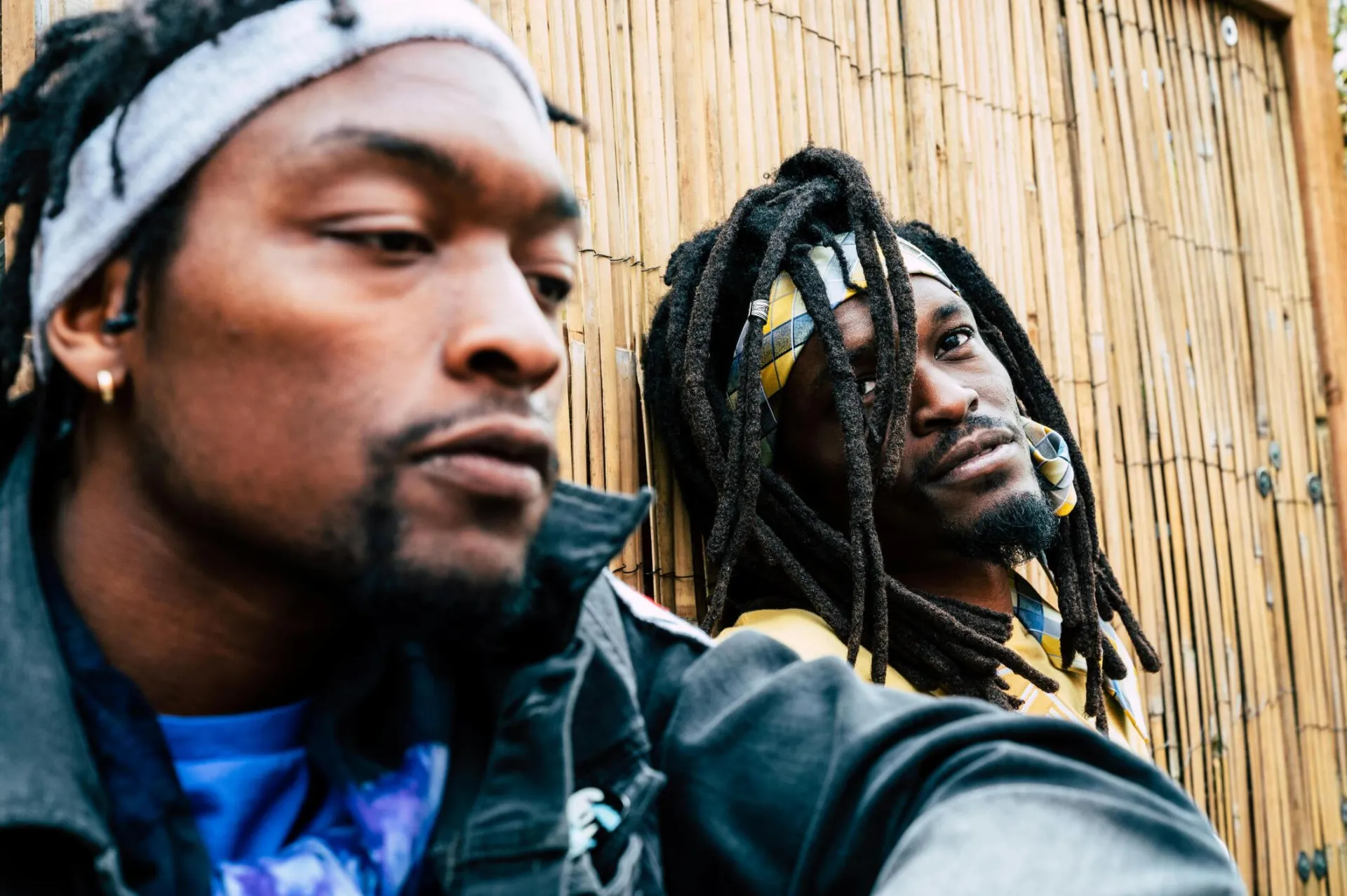 Two men with dreadlocks and headbands lean against a bamboo wall outdoors, captured in an urban setting.