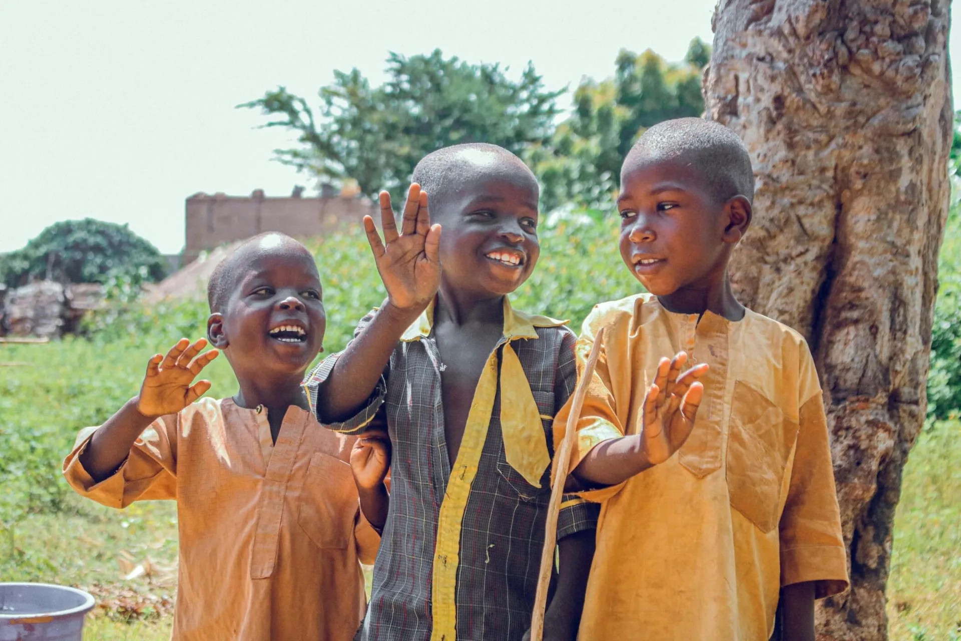 Three happy children waving outside in a vibrant Nigerian village setting.