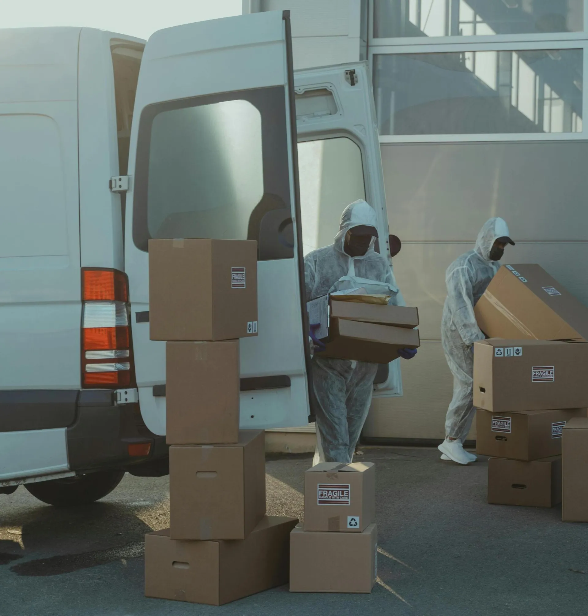 Two people unloading cardboard boxes from a van wearing protective suits, ensuring safety in package delivery.