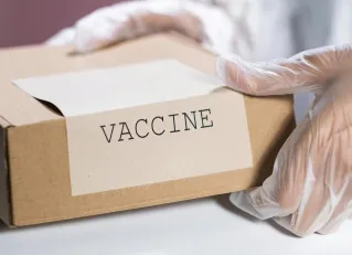 Gloved hands holding a labeled vaccine box in a clinical setting.