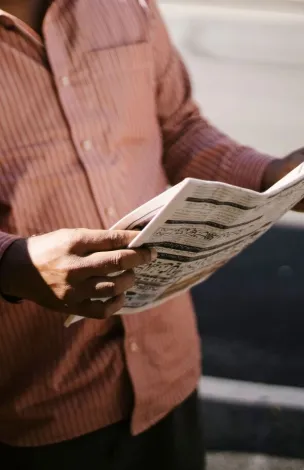 Crop anonymous ethnic cab driver in ornamental shirt standing with newspaper near transport in sunlight