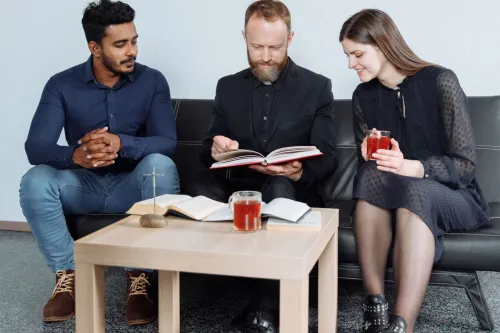 Three adults sitting indoors discussing religious text with open books and drinks.