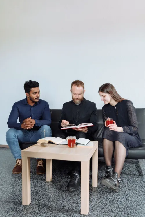 Three adults sitting indoors discussing religious text with open books and drinks.