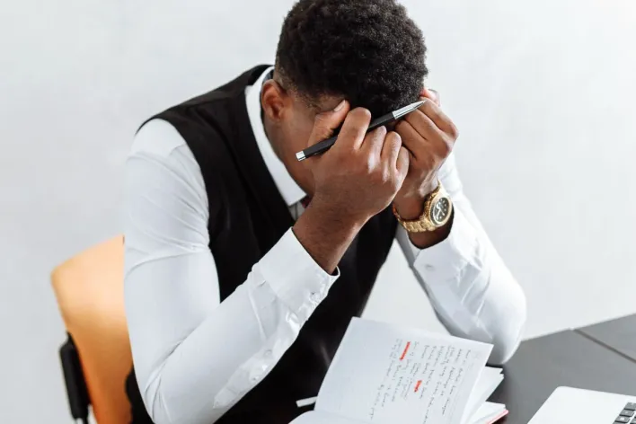 A businessman overwhelmed working indoors with a notebook, laptop, and hourglass.