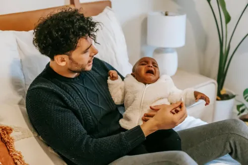 High angle of young ethnic father in casual clothes sitting on soft bed and hugging adorable crying African American baby during weekend at home