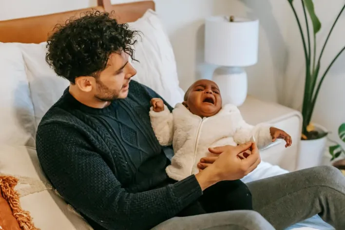 High angle of young ethnic father in casual clothes sitting on soft bed and hugging adorable crying African American baby during weekend at home