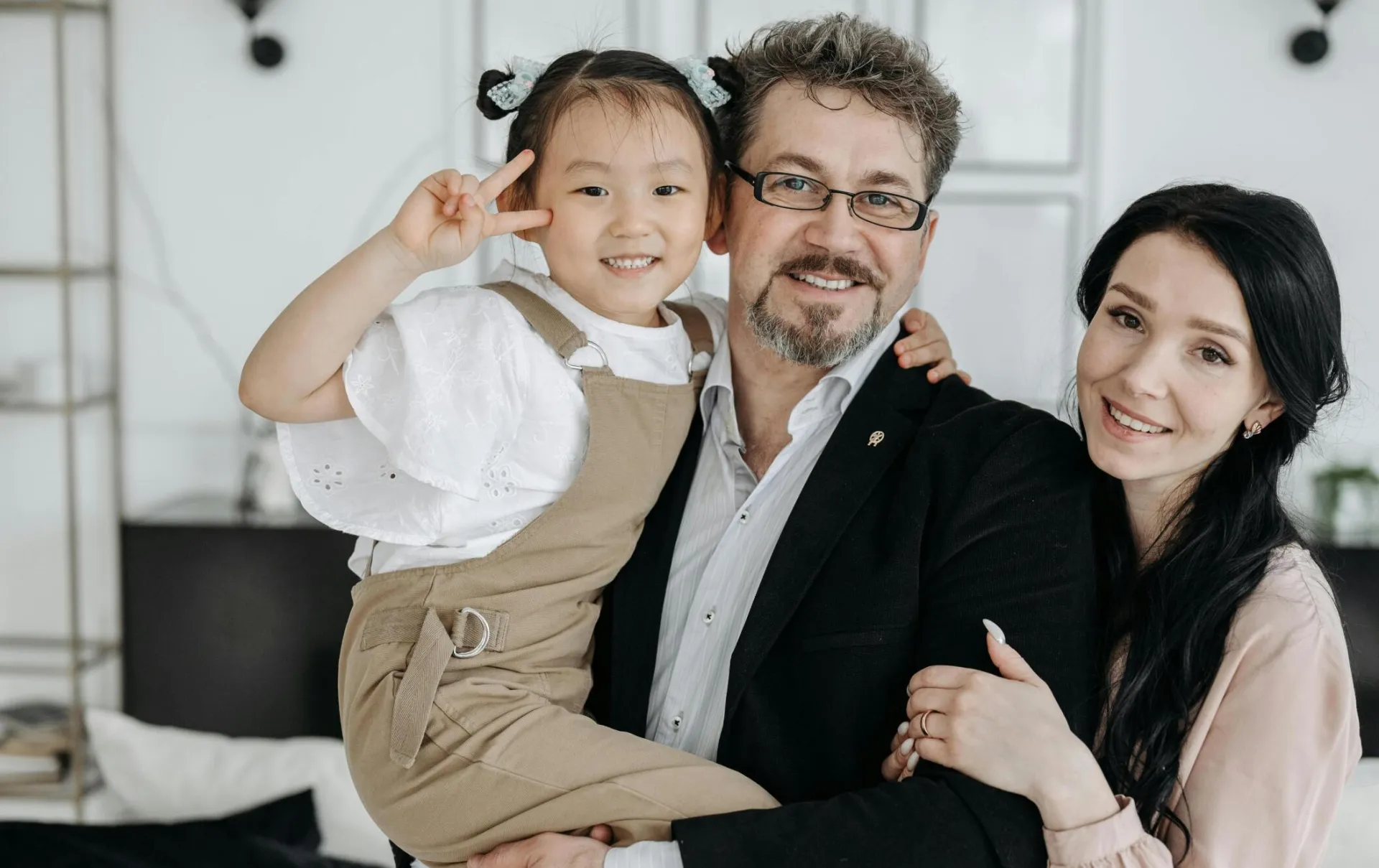 A joyful multicultural family portrait of parents and their adopted daughter smiling indoors.
