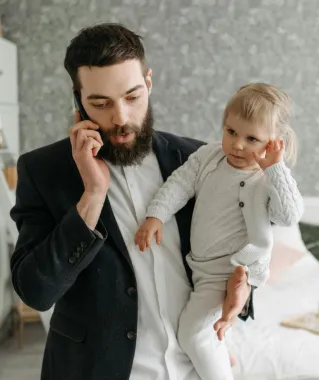 A bearded father talks on the phone while holding his baby in a cozy bedroom setting.