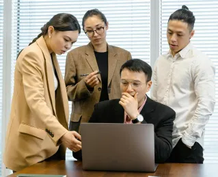 A group of colleagues engaged in a meeting, discussing around a laptop in a modern office.