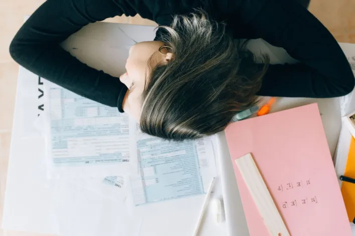 Exhausted woman resting her head on a desk cluttered with papers and a ruler, showing workplace fatigue.