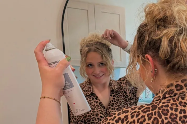 Woman styling her hair in front of bathroom mirror wearing a white shirt.