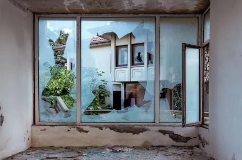 A shattered window revealing an abandoned building and greenery outside.