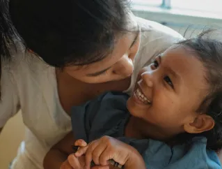High angle of cute little Asian girl smiling while sitting on knees of crop positive young mother pouting lips