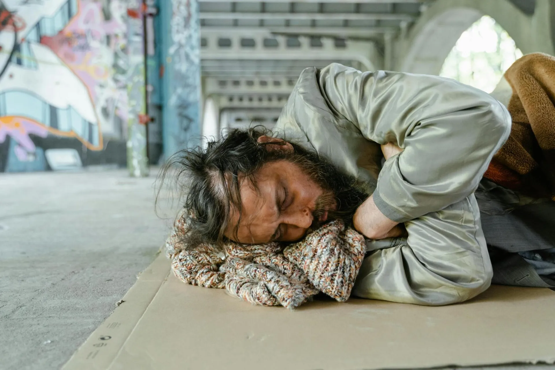 A homeless man sleeping on cardboard under a bridge, highlighting urban poverty.