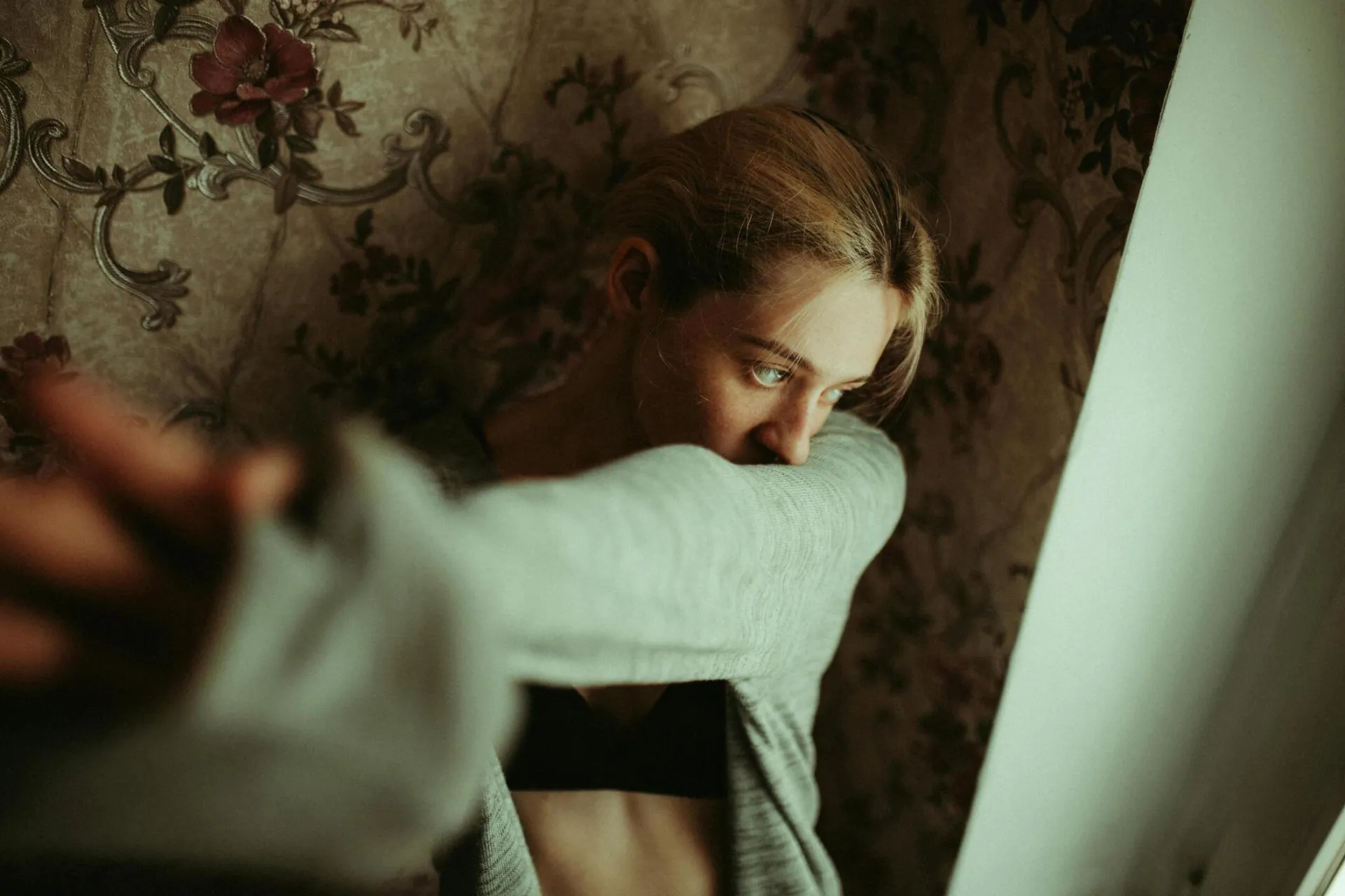 Young woman gazing thoughtfully out of window in room with floral wallpaper, creating a vintage and introspective mood.