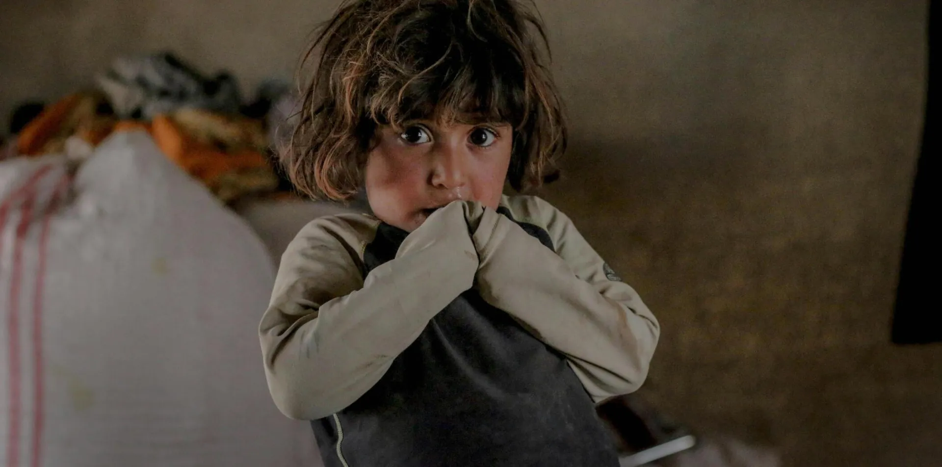 Little refugee child with dirty hair covering mouth with hands and looking at camera while standing in shabby room inside