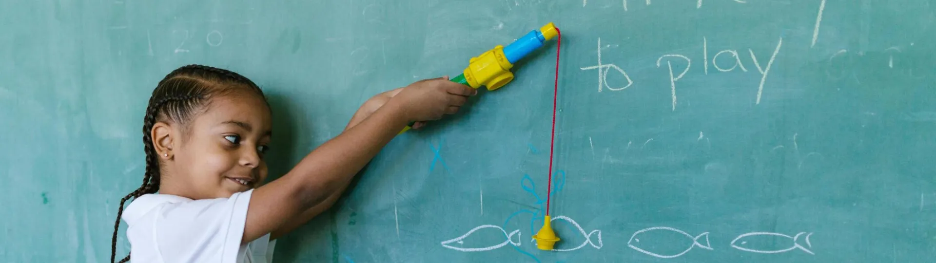 A young boy with braided hair playing with a toy fishing rod by a blackboard in a classroom setting.