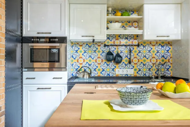 Wooden table with dishware and fresh fruits placed near white cupboards and modern kitchenware with utensils in light colorful kitchen at home