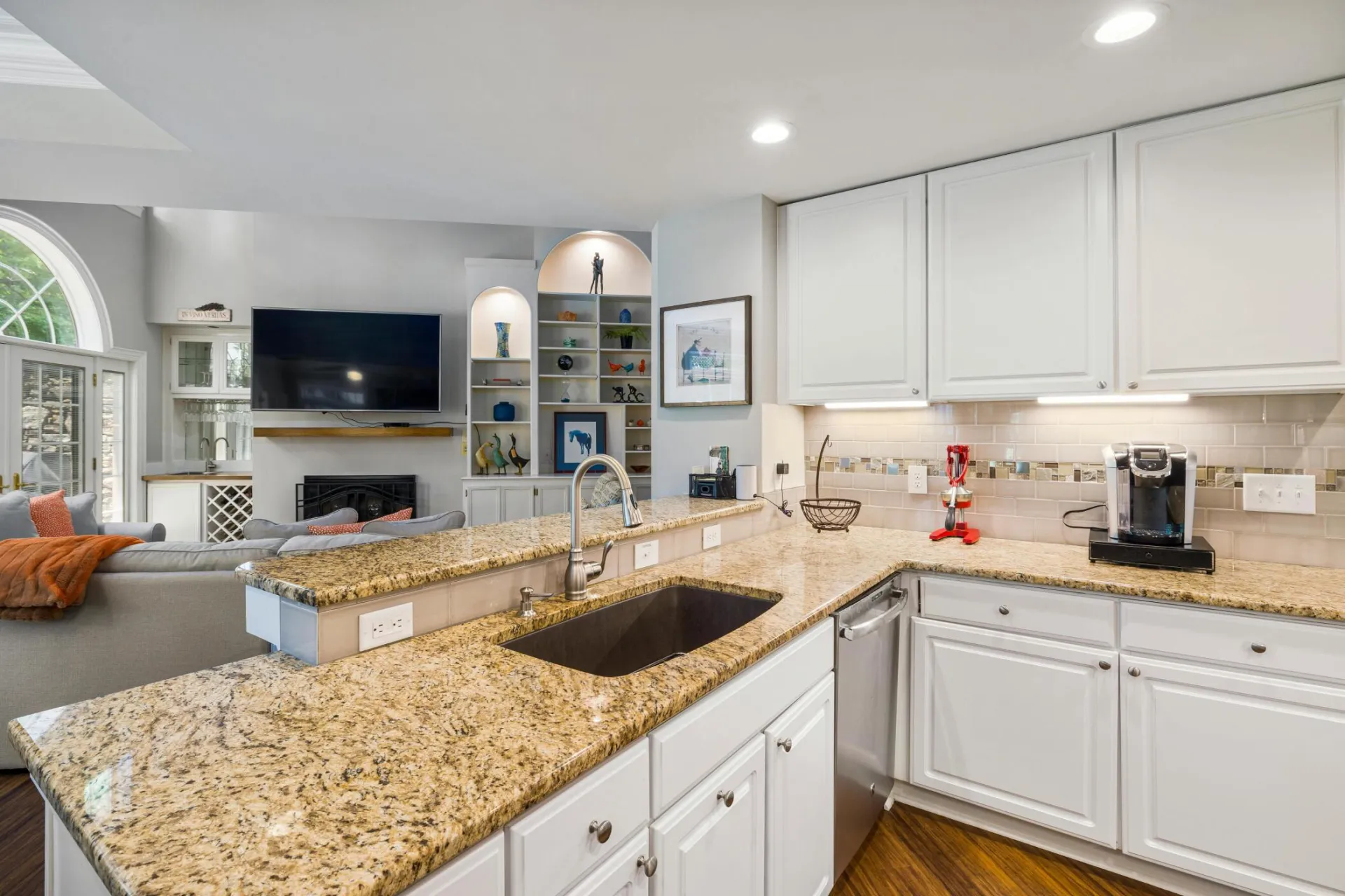 A modern kitchen with granite countertops opens into a stylish living room, featuring elegant furniture and built-in shelves.
