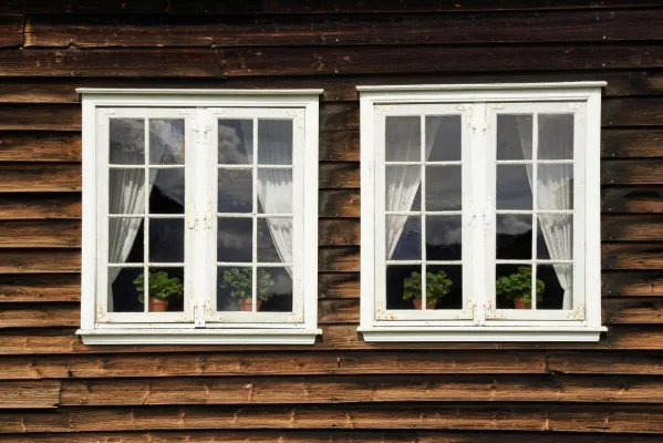 Close-up of two white framed windows with curtains against a rustic wooden house exterior.