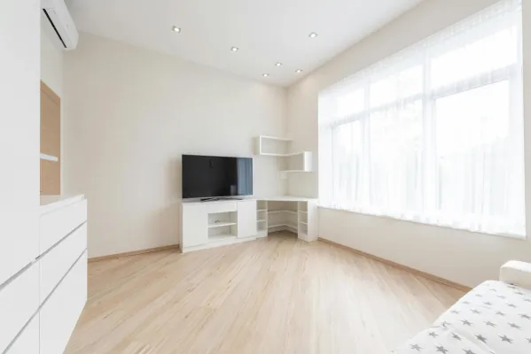 Modern TV set on cupboard placed near empty table in spacious room with big window and white cabinets in apartment