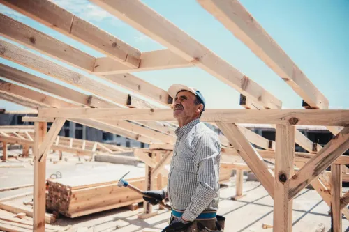 A construction worker surveys a wooden roof frame build on a sunny day.