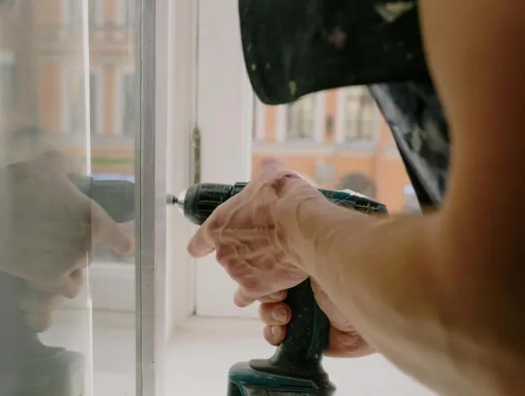 Unrecognizable professional male worker assembling white window with cordless screwdriver with drill near windowsill with reflection on glass during repair works in apartment