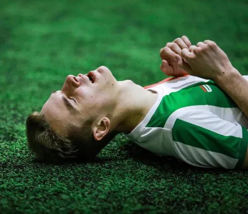 Young male soccer player in green jersey lying on grass, eyes closed, exhausted.