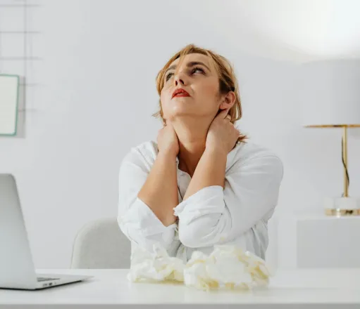 A stressed woman in white long sleeves sits at a desk with a laptop, looking upwards in an office.