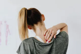 Back view of slim blond female in casual wear with manicure indicating pain spot on back while sitting in front of white wall with paper drawing in clinic