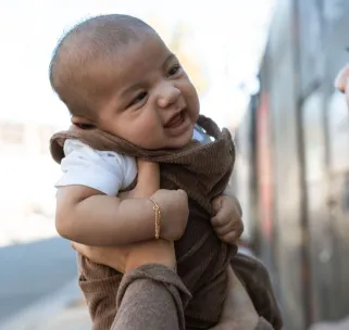Cute baby smiling while being held outdoors, wearing a brown outfit with a blurred background.