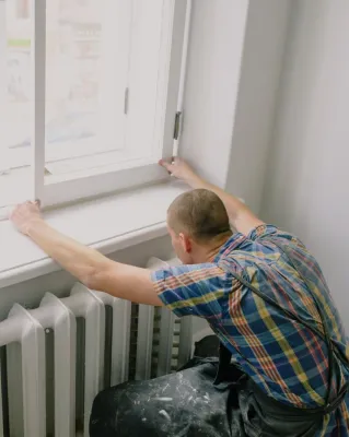 Back view of unrecognizable fitter in dirty apron installing window above radiator while doing home renovation