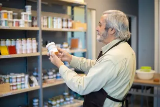 Senior man in apron organizing paint jars on a store shelf indoors.