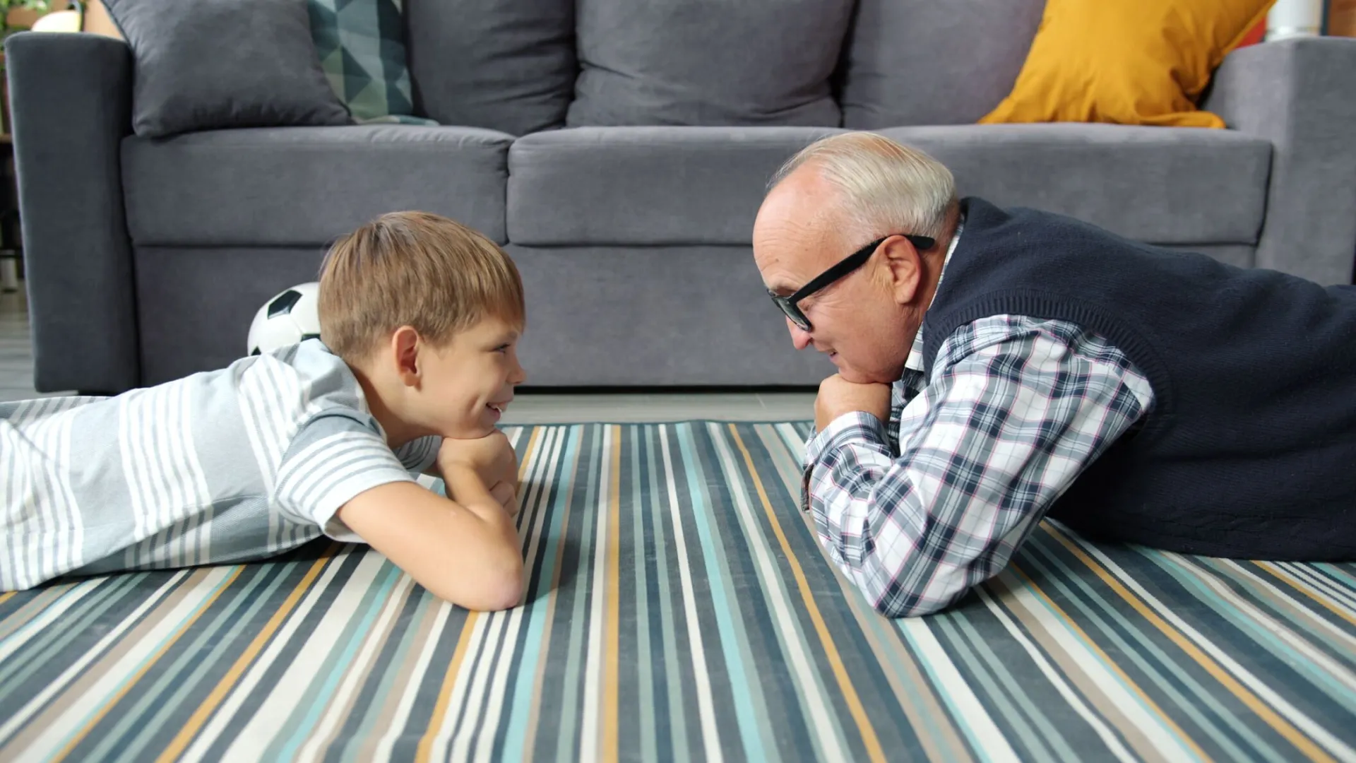 A charming connection between grandfather and grandson lying face-to-face indoors on a colorful carpet.