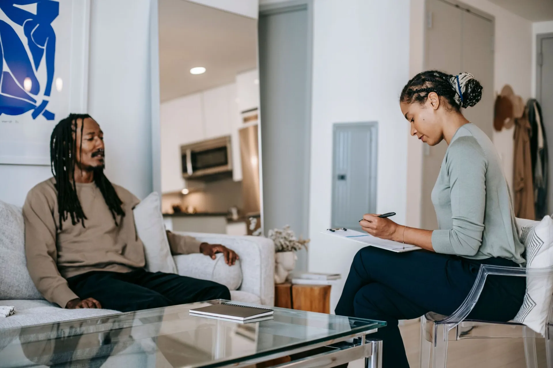 Focused African American female therapist writing on clipboard while sitting in front of upset black male client during conversation on blurred background