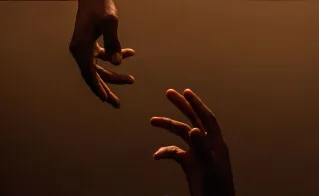 Two hands reaching towards each other against a dark, warm background, symbolizing connection.