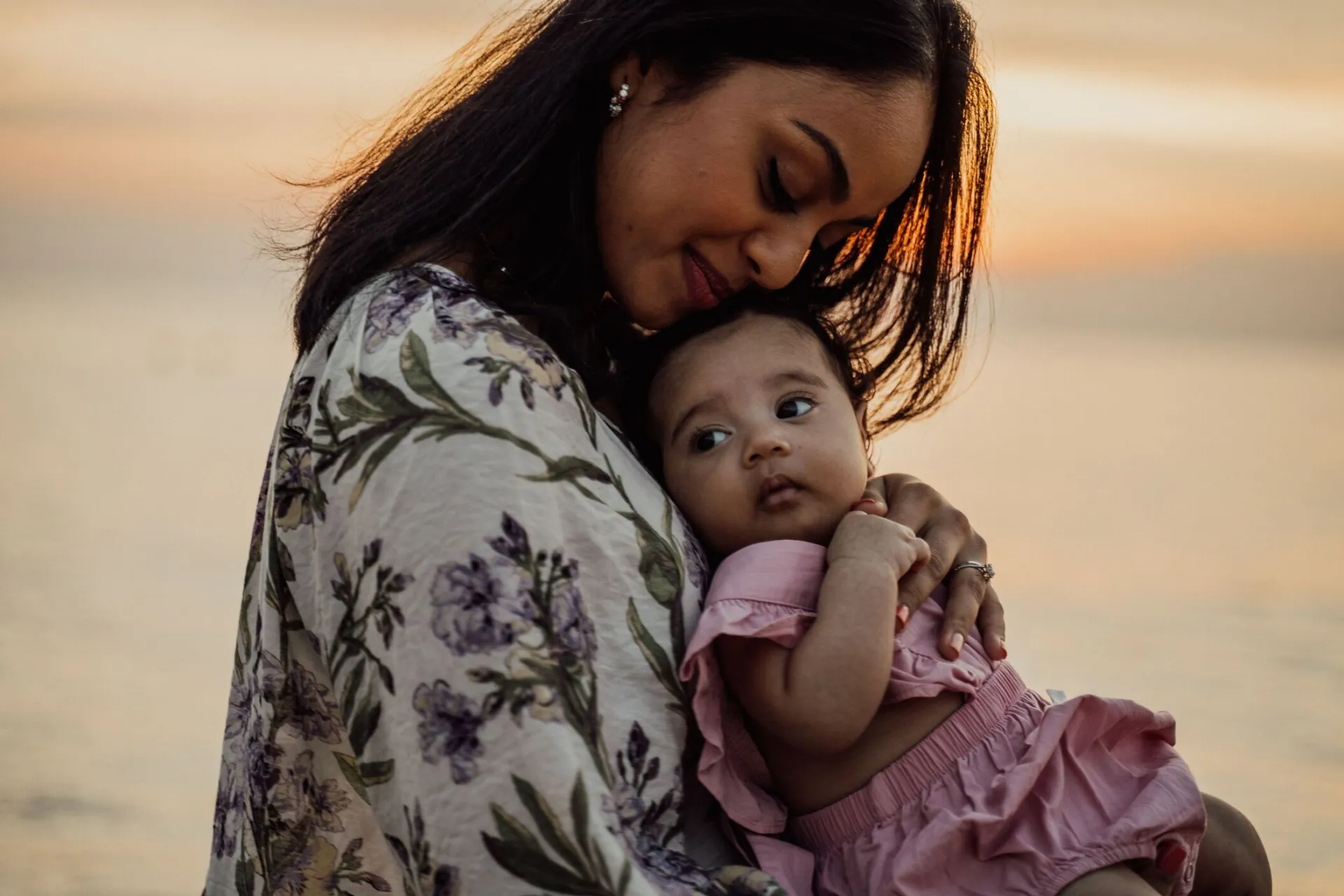 A loving mother holding her baby at the beach during a beautiful sunset.
