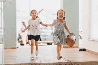 Two smiling girls running and holding hands indoors with a paper bag, conveying happiness and friendship.