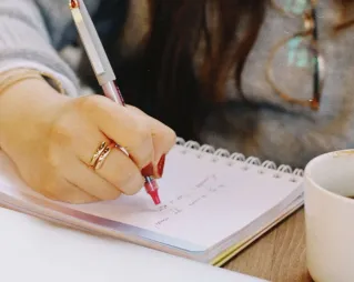 Close-up of a woman's hand writing notes in a notebook with a cup of coffee nearby.