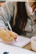 Close-up of a woman's hand writing notes in a notebook with a cup of coffee nearby.